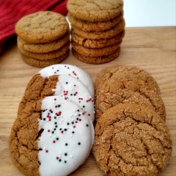 gingersnap cookies stacked on a board, some of them have been half-dipped in white chocolate and have sprinkles for Christmas.