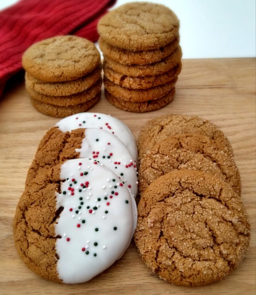 gingersnap cookies stacked on a board, some of them have been half-dipped in white chocolate and have sprinkles for Christmas.