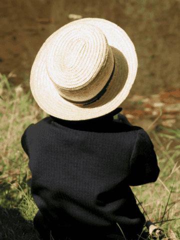 an Amish boy with a hat sitting in the grass by the water with his back turned.