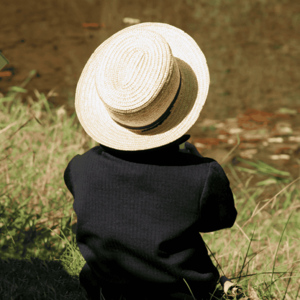 an Amish boy with a hat sitting in the grass by the water with his back turned.