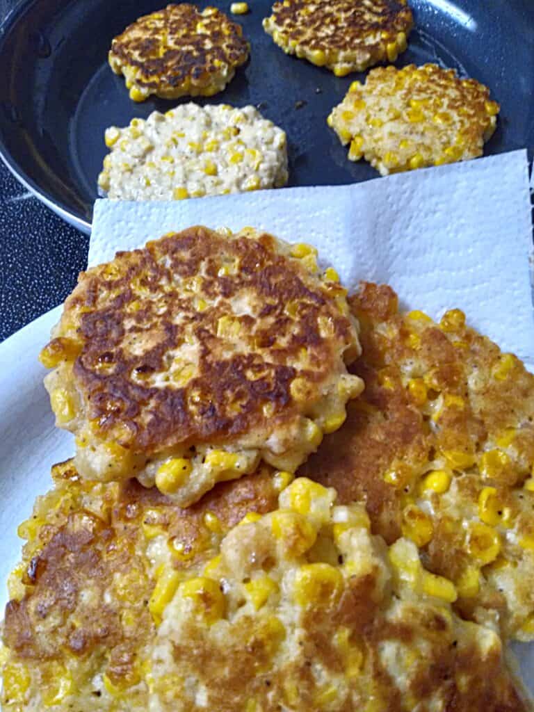 frying corn fritters in a skillet, some are on a paper towel-lined plate.