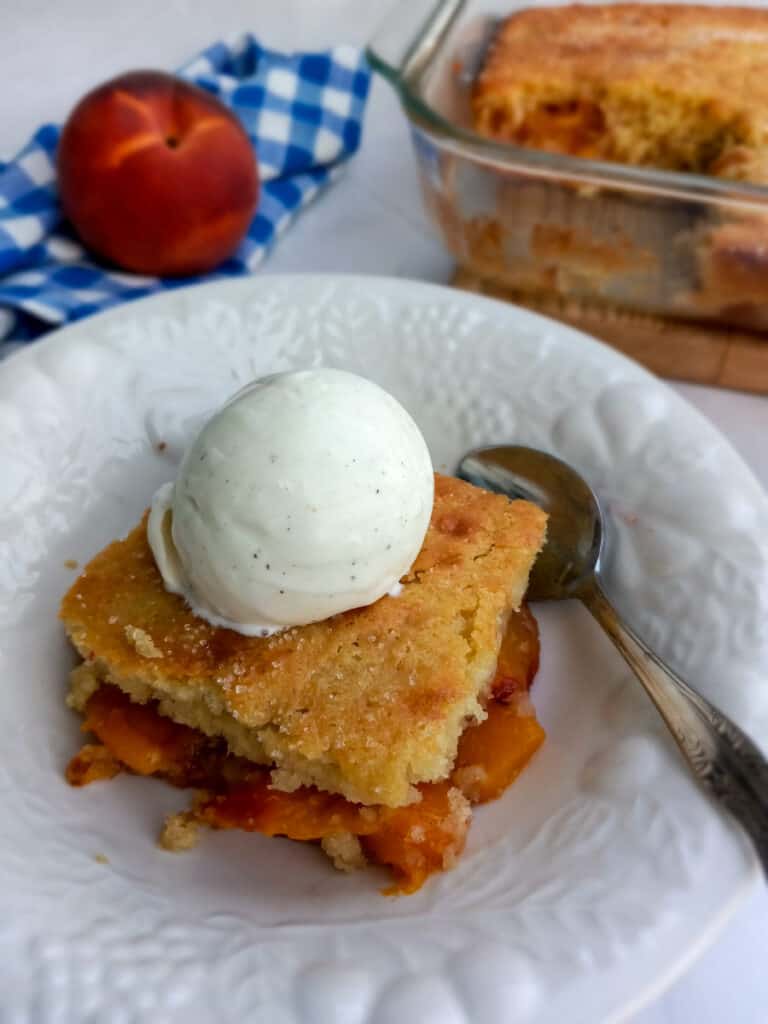  a dish with cobbler and ice cream and a 9x13" pan of cobbler, a blue towel, and a peach in the background
