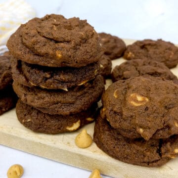 Amich chocolate cookies with peanut butter chips stacked on a board with peanut butter chips scattered around
