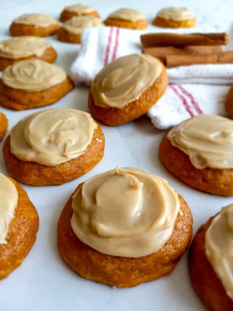 pumpkin cookies with caramel frosting scattered around, with a towel and cinnamon sticks for decor