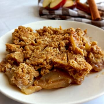 Amish apple goodie on a plate with a towel, apples, and cinnamon sticks in the background