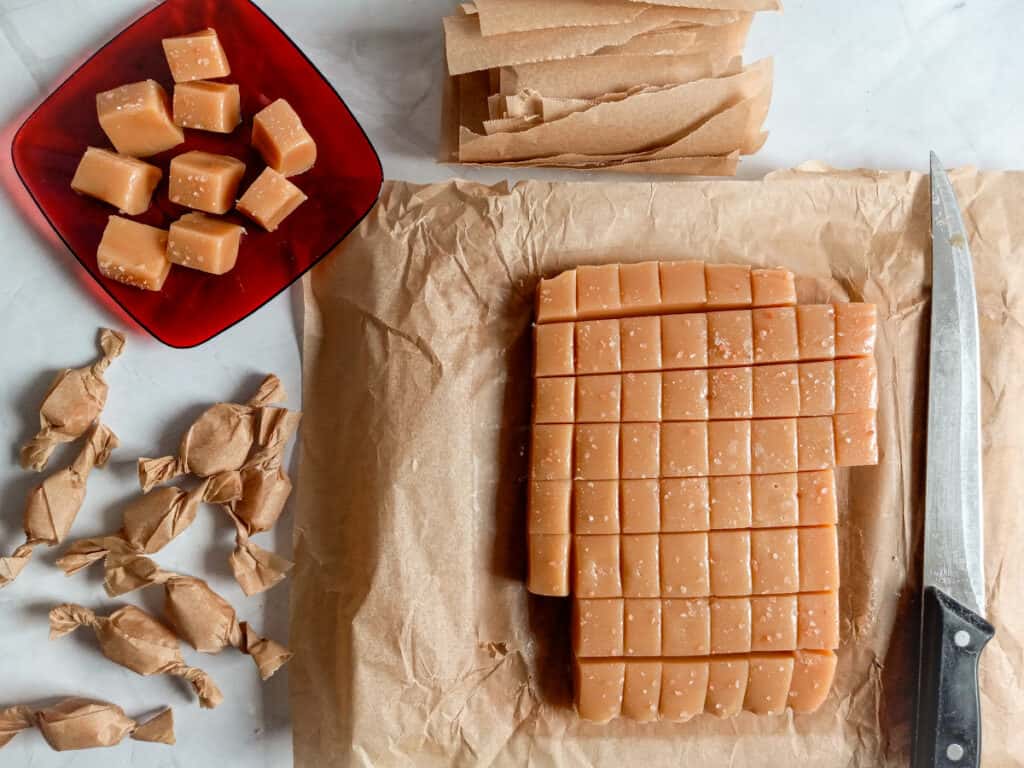 a few homemade caramels on a plate, a stack of parchment paper wrappers, a big piece of cut caramels on a board with a large knife, and some of the candies are wrapped individually.