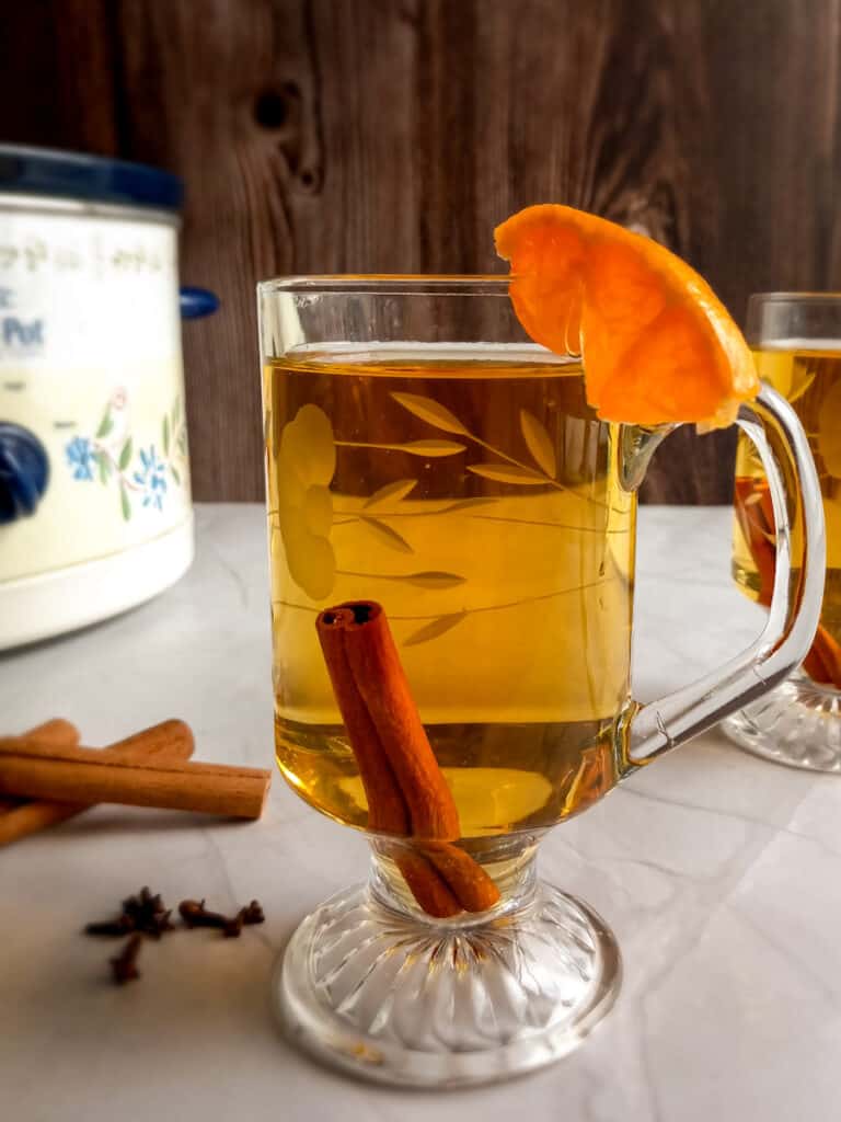 a cup of hot cider with a cinnamon stick, with the crockpot in the background.