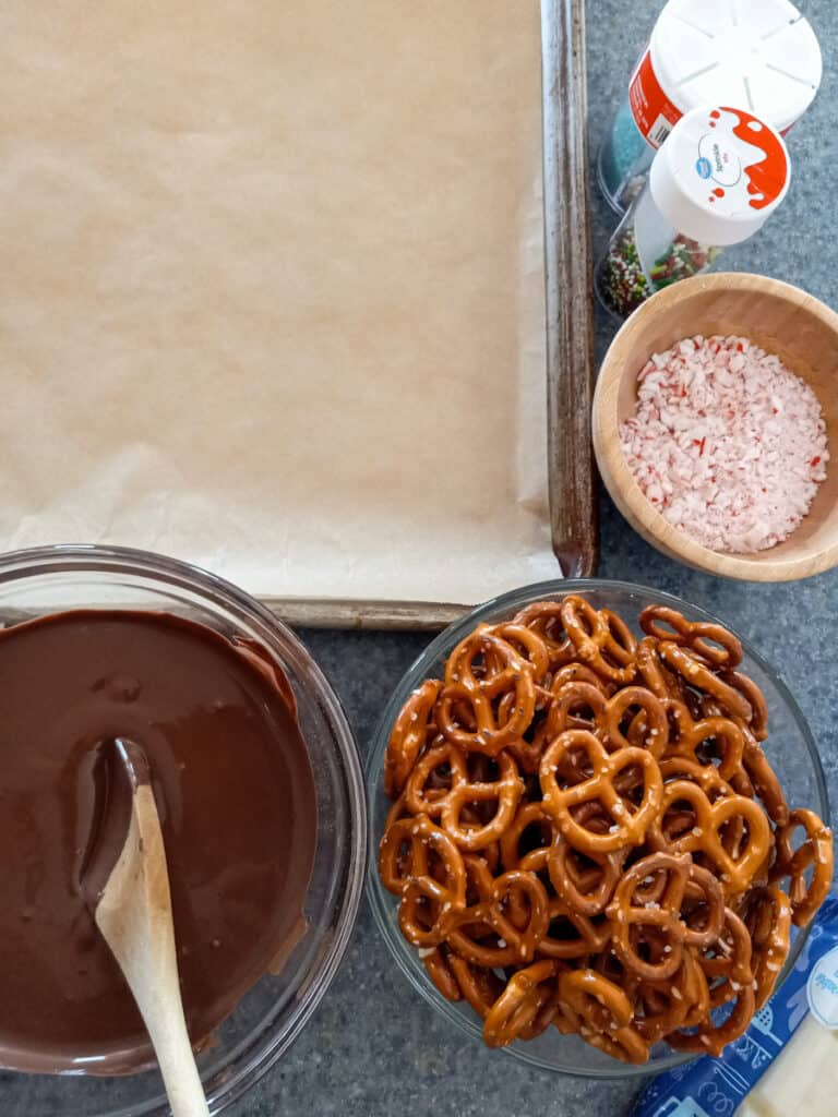 melted chocolate, pretzels in a bowl, parchment-lined tray, and sprinkles.
