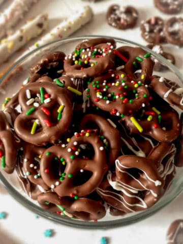 a clear glass bowl full of chocolate pretzel twists with sprinkles, pretzel rods in the background.