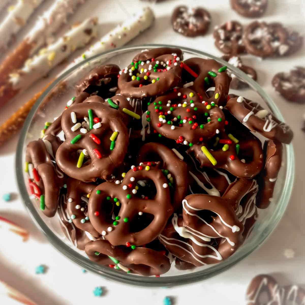 a clear glass bowl full of chocolate pretzel twists with sprinkles, pretzel rods in the background.
