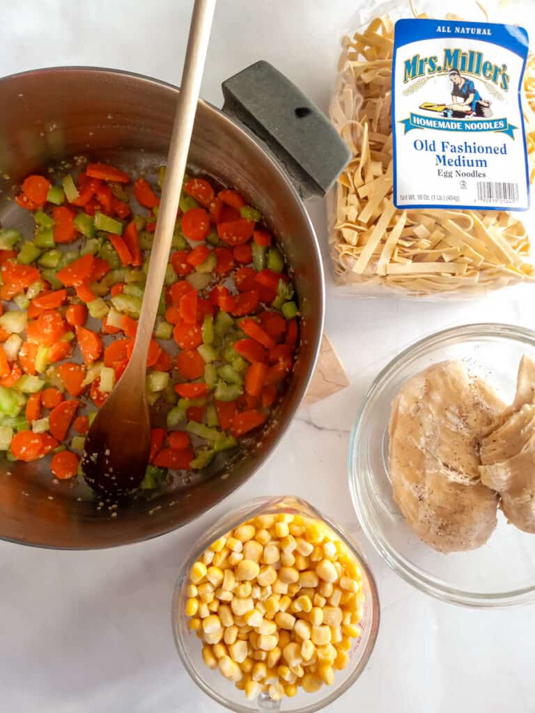 chopped carrots, celery, and garlic in a large pot, noodles, corn, and chicken in separate dishes.