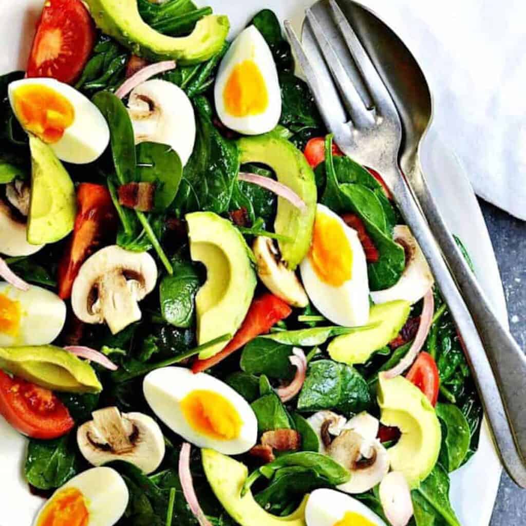 a plate fo spinach salad with boiled eggs, avocado, and mushrooms.
