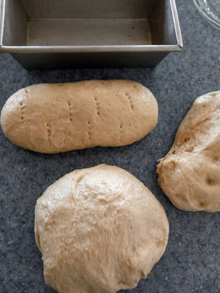 bread dough divided into three pieces, one loaf shaped and ready to put into pan.