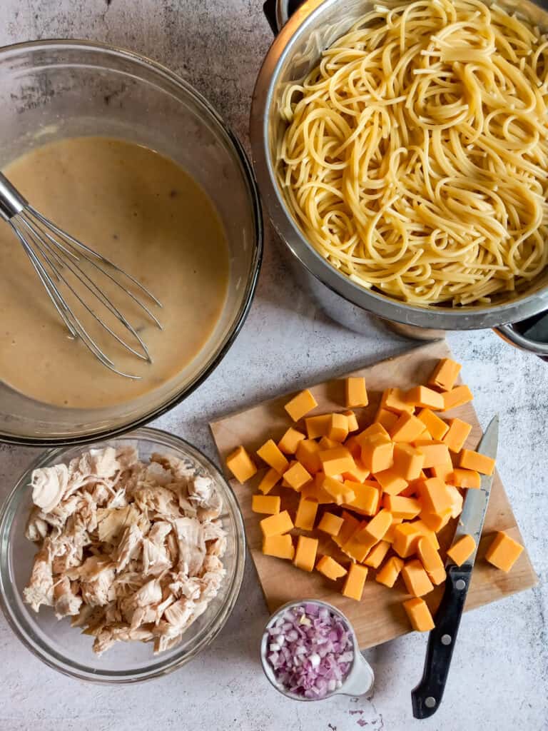 cooked spaghetti in a strainer, a bowl with the wet ingredients, a bowl of cooked chicken, cut cheese cubes on a board, and chopped onion in a small bowl.