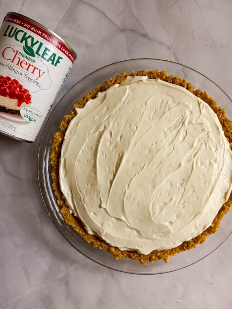graham crust filled with cheesecake filling and a can of cherry pie filling ready to go on the top.