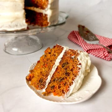a slice of Amish carrot cake with pineapple and pecans and cream cheese frosting, the remaining cake on a cake stand in the background.