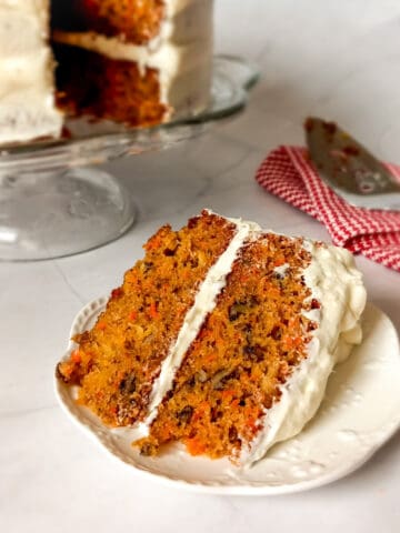 a slice of Amish carrot cake with pineapple and pecans and cream cheese frosting, the remaining cake on a cake stand in the background.