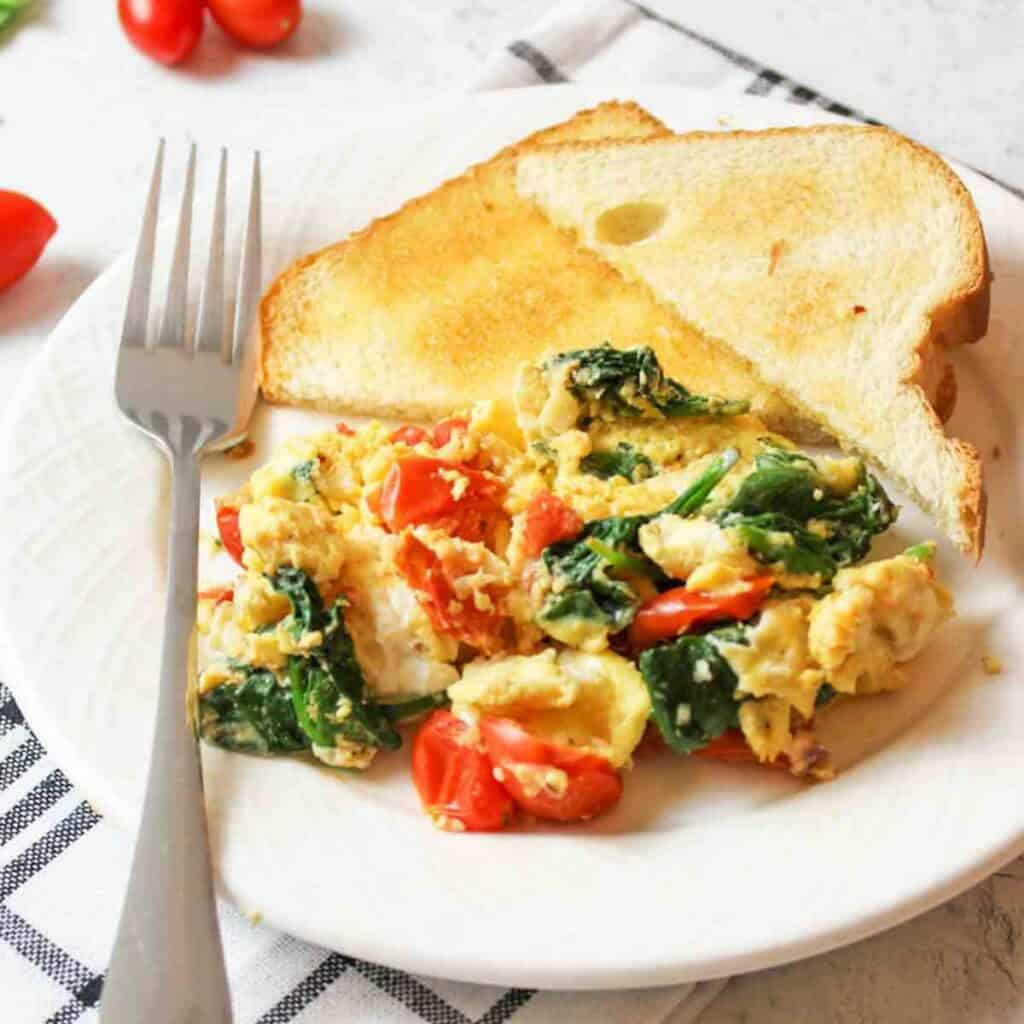 scrambled eggs with spinach and tomato on a plate with toast.