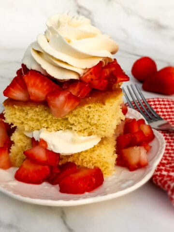 spongey strawberry shortcake cut in half and on a plate, topped with strawberries and whipped cream.