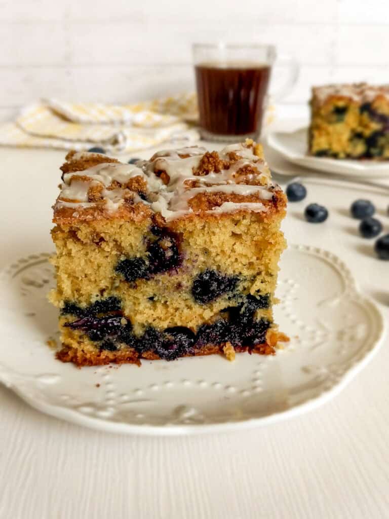 two slices of blueberry coffee cake on small plates with a cup of coffee in the background.