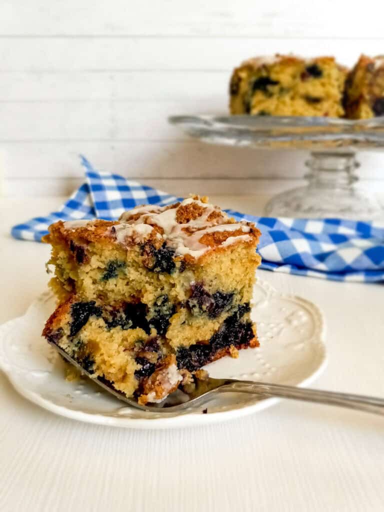 cutting into a slice of blueberry cake on a plate, and a cake stand with more cake in the background.