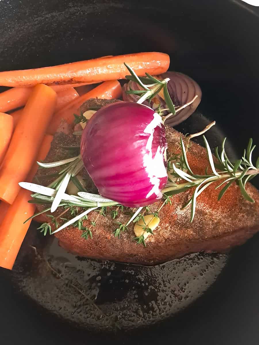 a roast in the Dutch oven topped with seasonings, garlic, and onion, and carrots on the side.