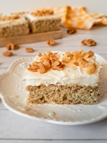 a banana cream cheese cake bar on a small plate, with more in the background, and walnuts scattered around.