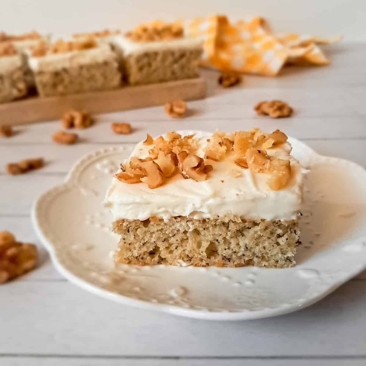 a banana cream cheese cake bar on a small plate, with more in the background, and walnuts scattered around.