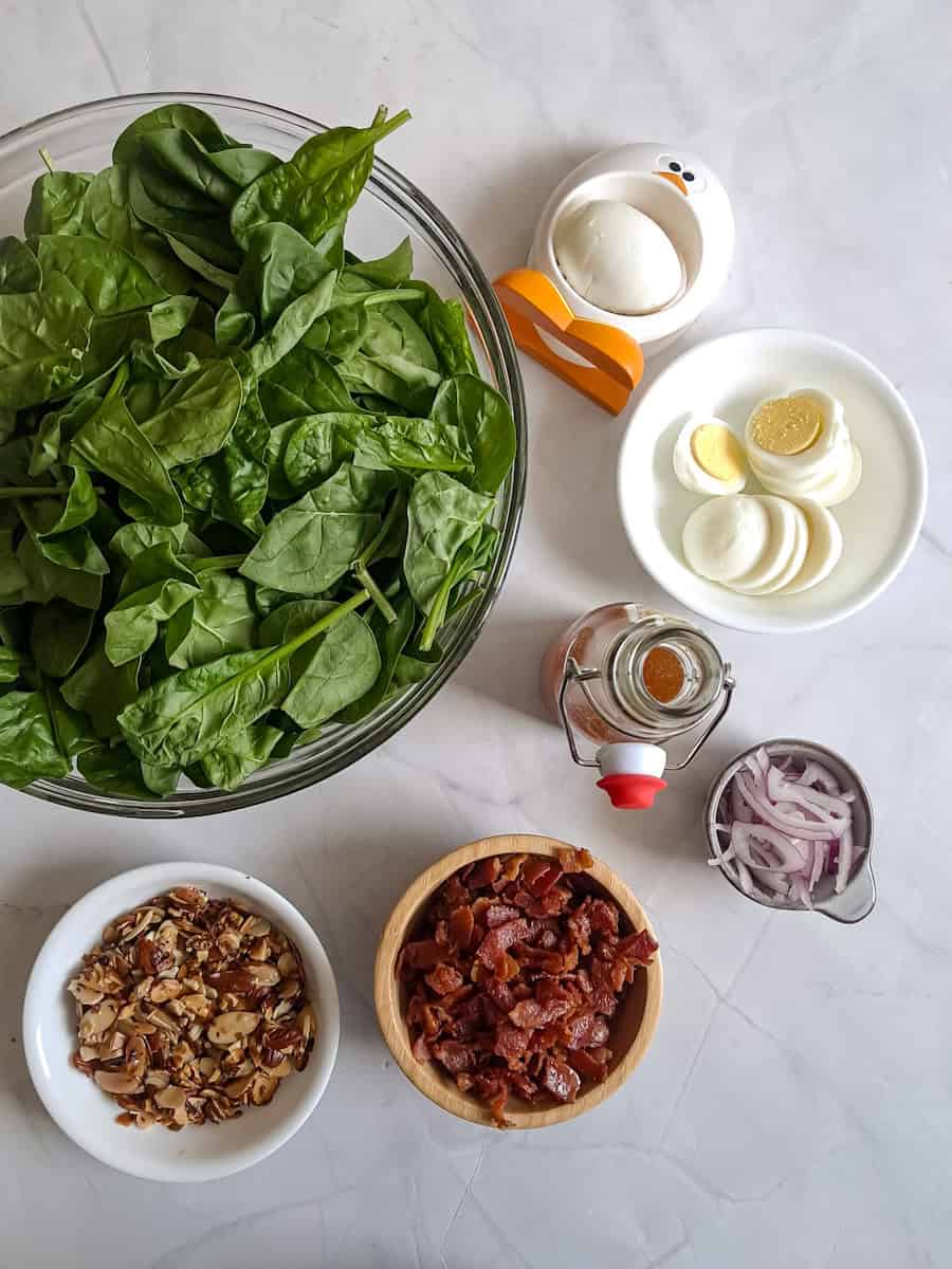 a big bowl of spinach, and small bowls of toasted almonds, fried bacon, boiled eggs, and sliced onions.