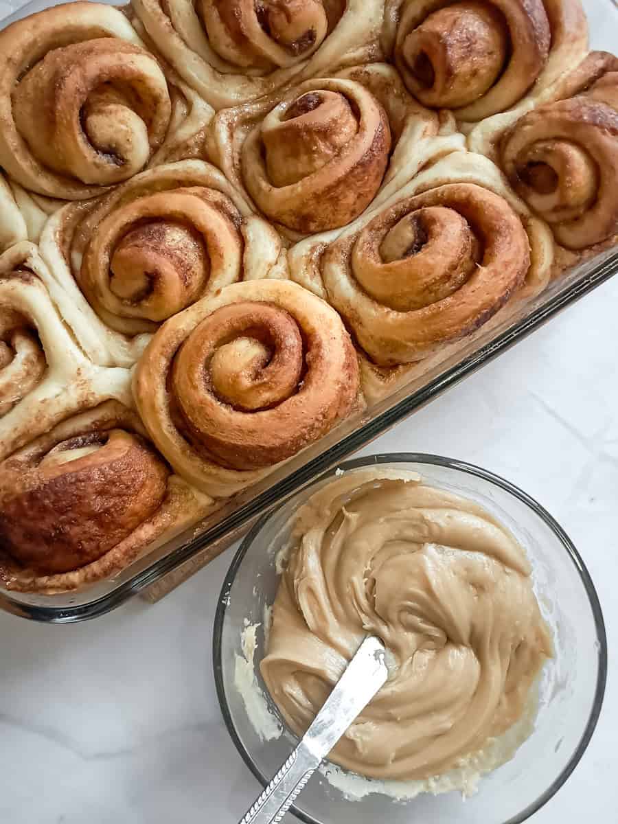 baked cinnamon rolls are ready to frost with a bowl of caramel frosting.