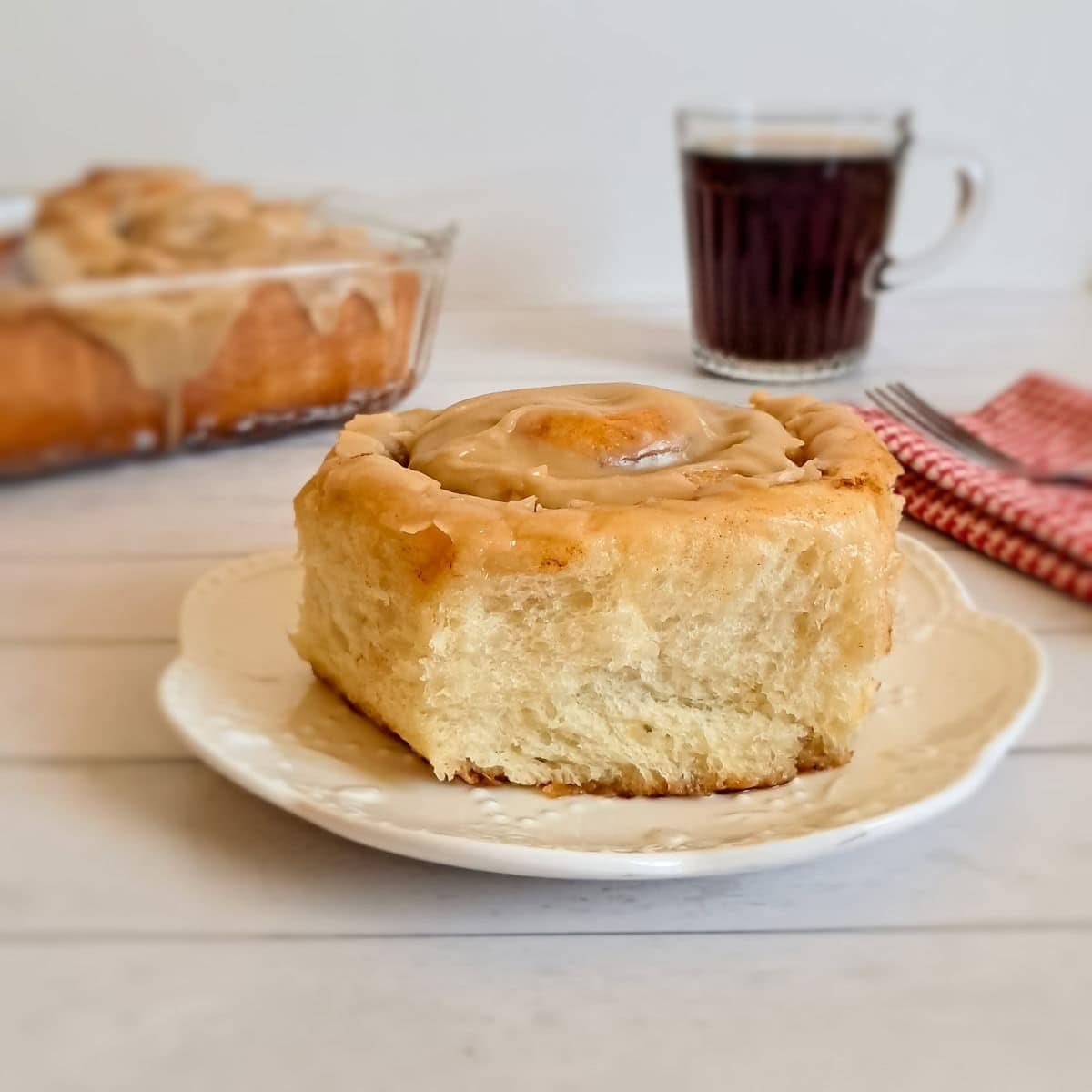 cinnamon roll on a plate and remaining pan of rolls with coffee in the background.