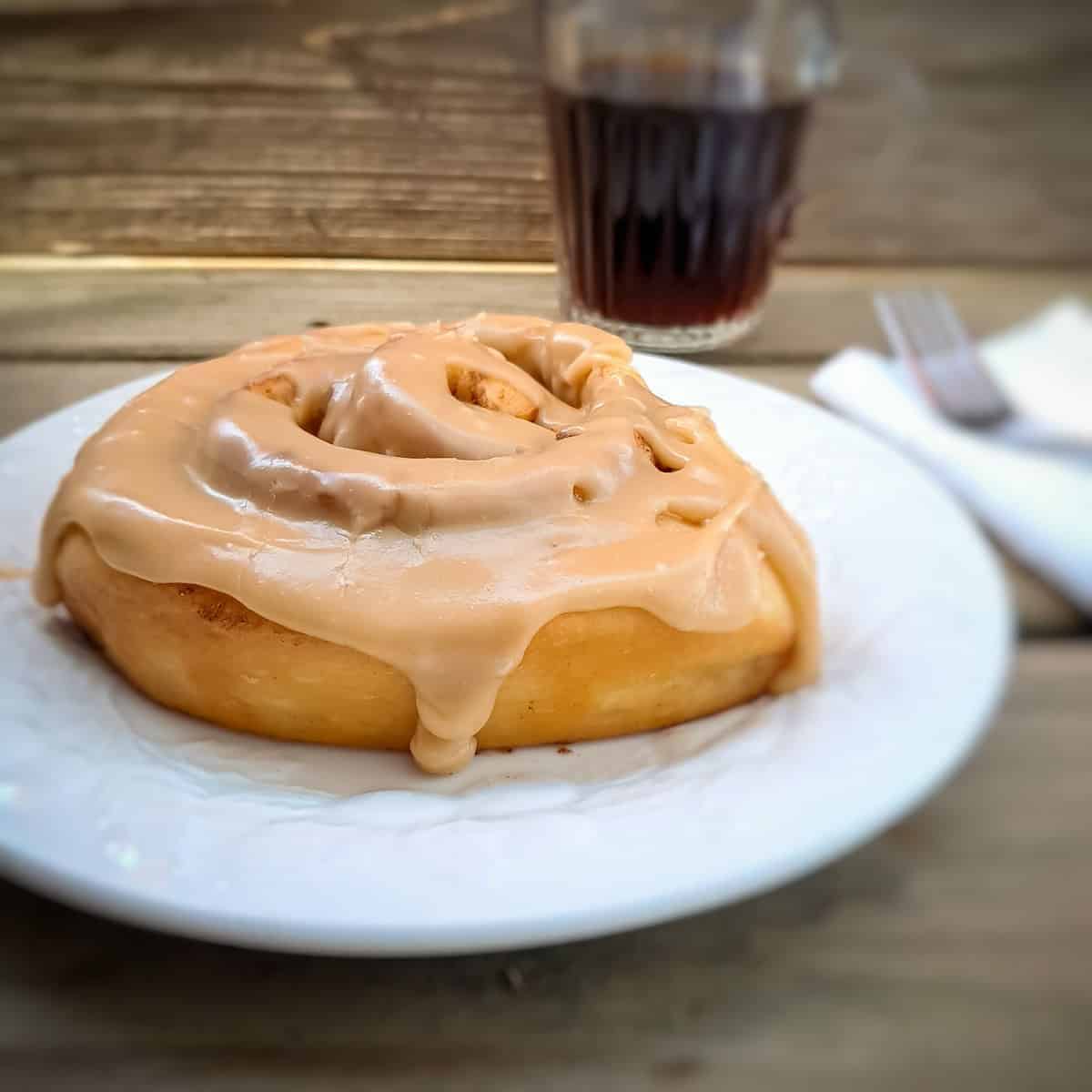 Aish cinnamon roll covered in caramel frosting on a plate with a cup of coffee on the side.