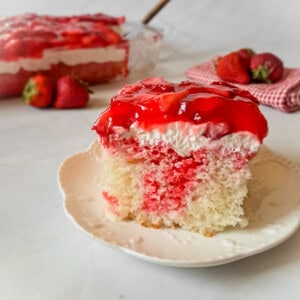 a slice of Amish fruit jello cake on a plate, strawberries and the remaining pan of dessert in the background.