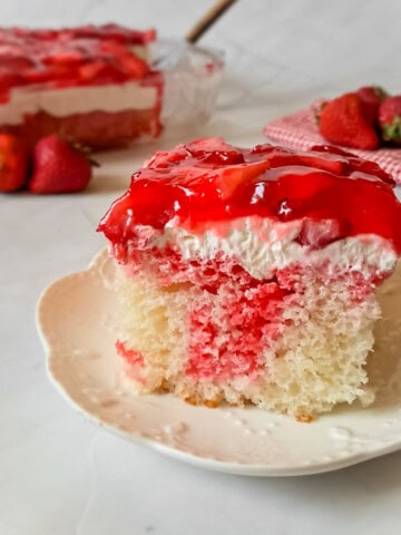 a slice of Amish fruit jello cake on a plate, strawberries and the remaining pan of dessert in the background.