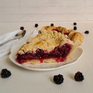 slice of blackberry pie on a small white plate, blackberries scattered around.