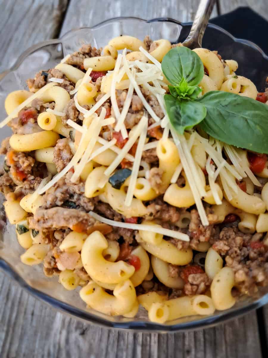 a glass bowl of Amish-style goulash with parmesan shreds and fresh basil on the top.