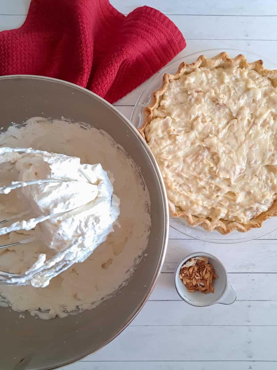 coconut cream filling in a pie crust, whipped cream is in a large bowl, and toasted coconut flakes, all ready to top the pie.