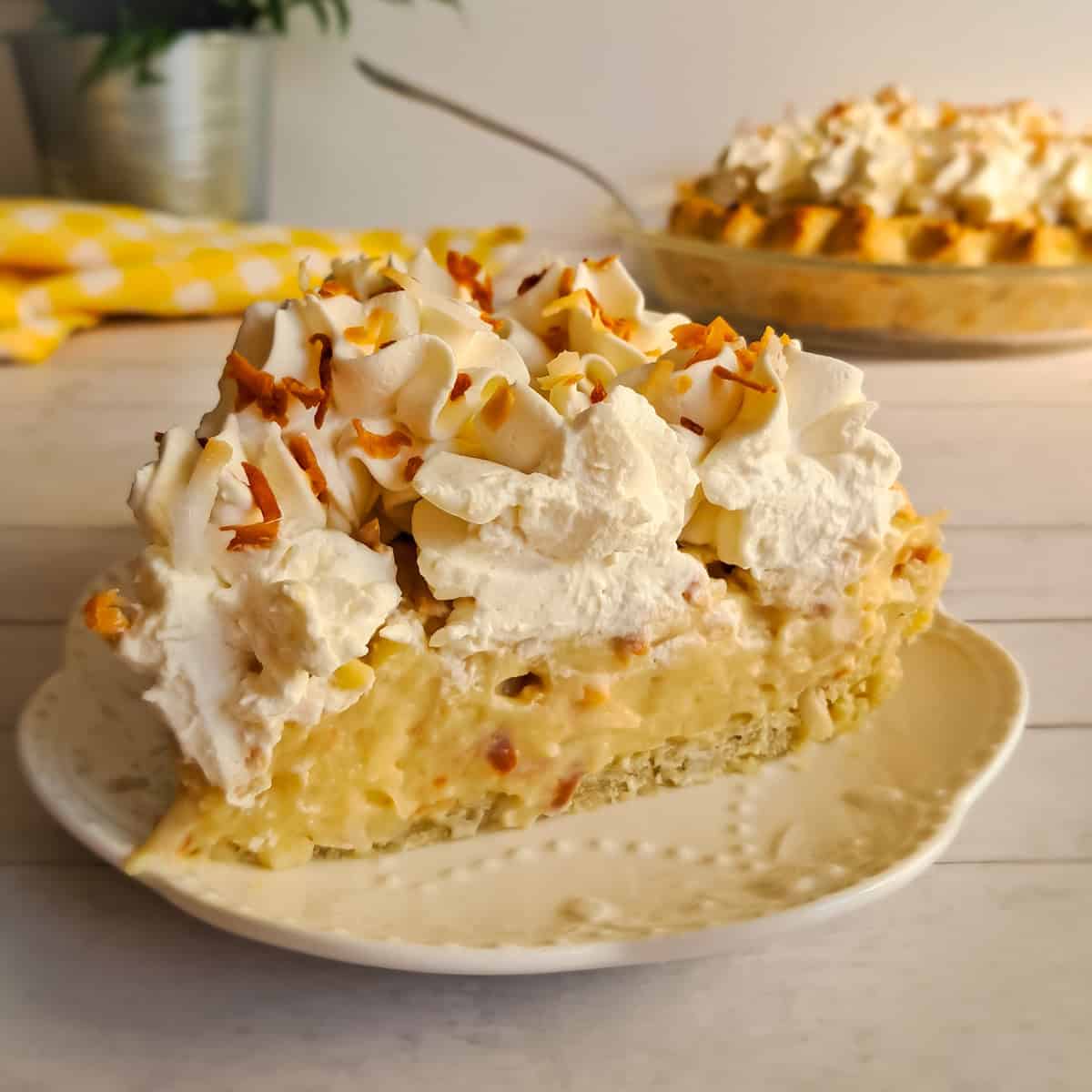 a slice of classic coconut cream pie on a plate with the remaining pie in the background.