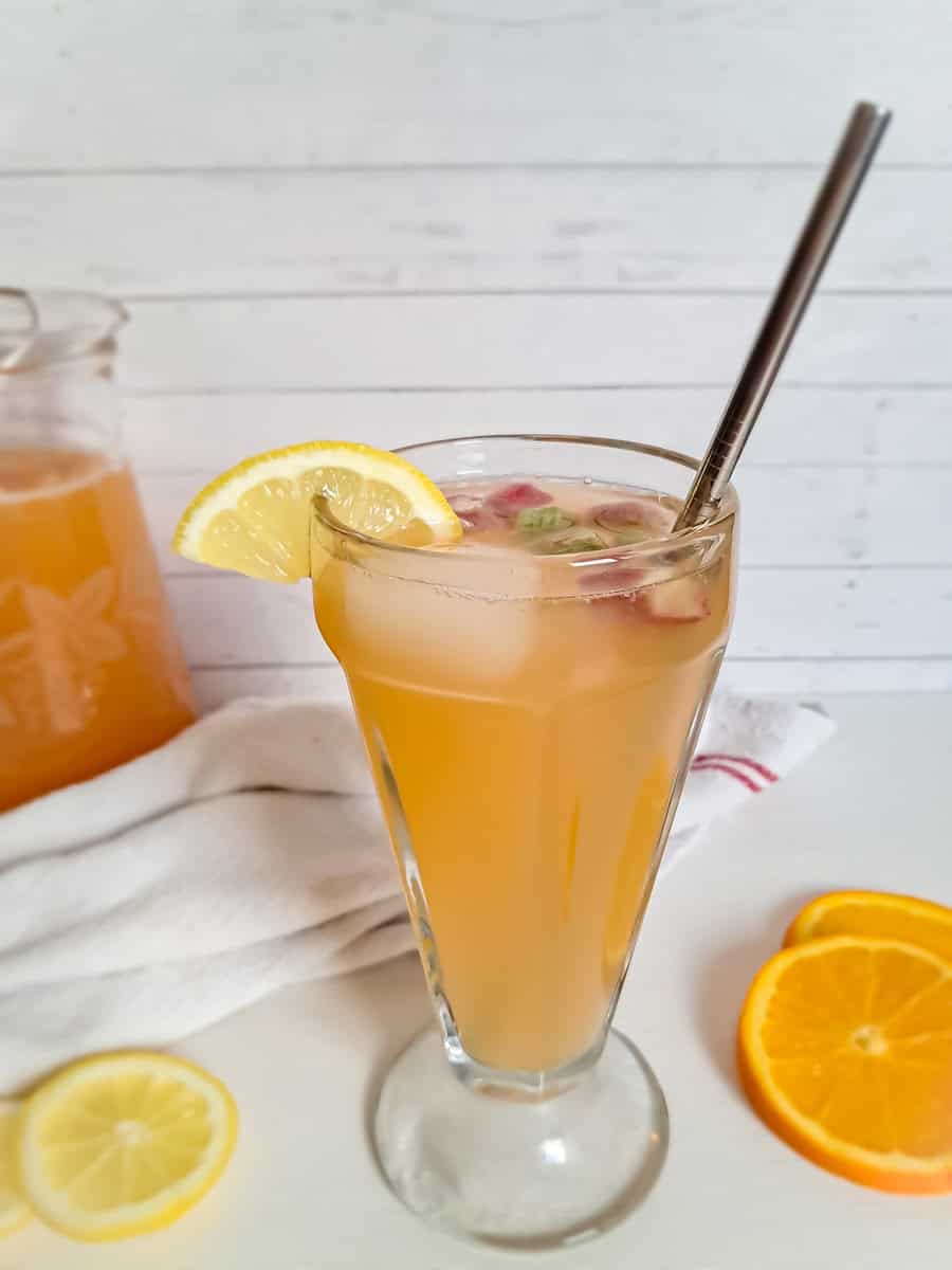 a pitcher and tall glass of rhubarb punch with lemon slice, ice, and rhubarb pieces.