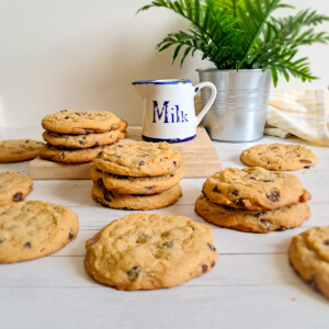 A bunch of Amish peanut butter chocolate chip cookies stacked and scattered around with a milk pitcher and plant in the background.
