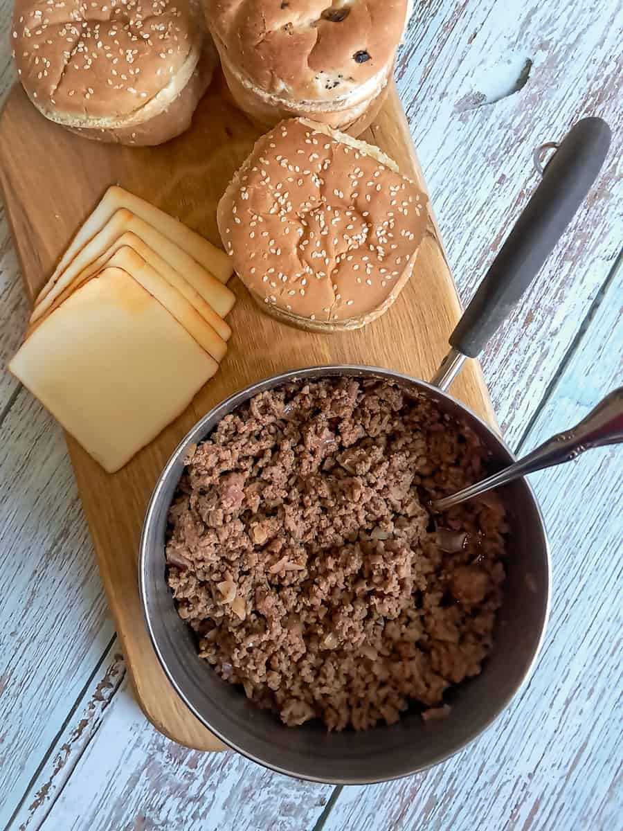 hamburger buns, cheese slices, and a saucepan full of sloppy joe meat on a board.