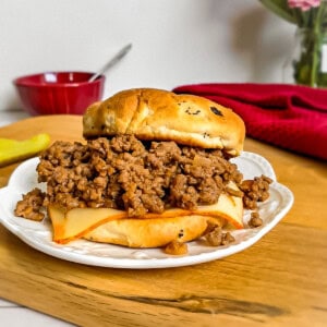a sloppy joe sandwich on a plate with a pickle and a red bowl of coleslaw on the side.