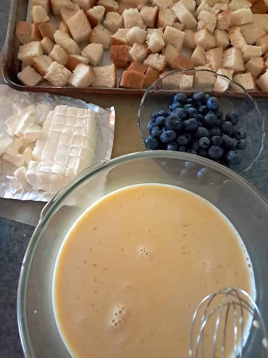 bread cubes on a tray, blueberries in a bowl, egg mixture in another bowl, and cream cheese cubes - ready to assemble.