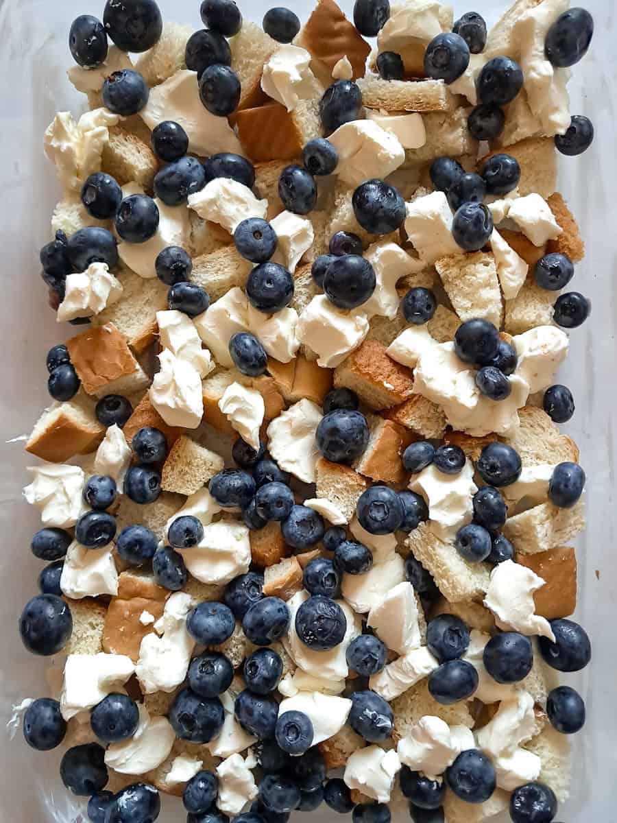 some of the bread cubes in the bottom of a 9x13" baking dish, cream cheese cubes, and blueberries scattered over the top.