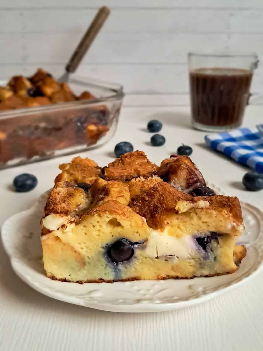 a slice of blueberry cream cheese french toast on a small plate, the remaining dish and a cup of coffee in the background.